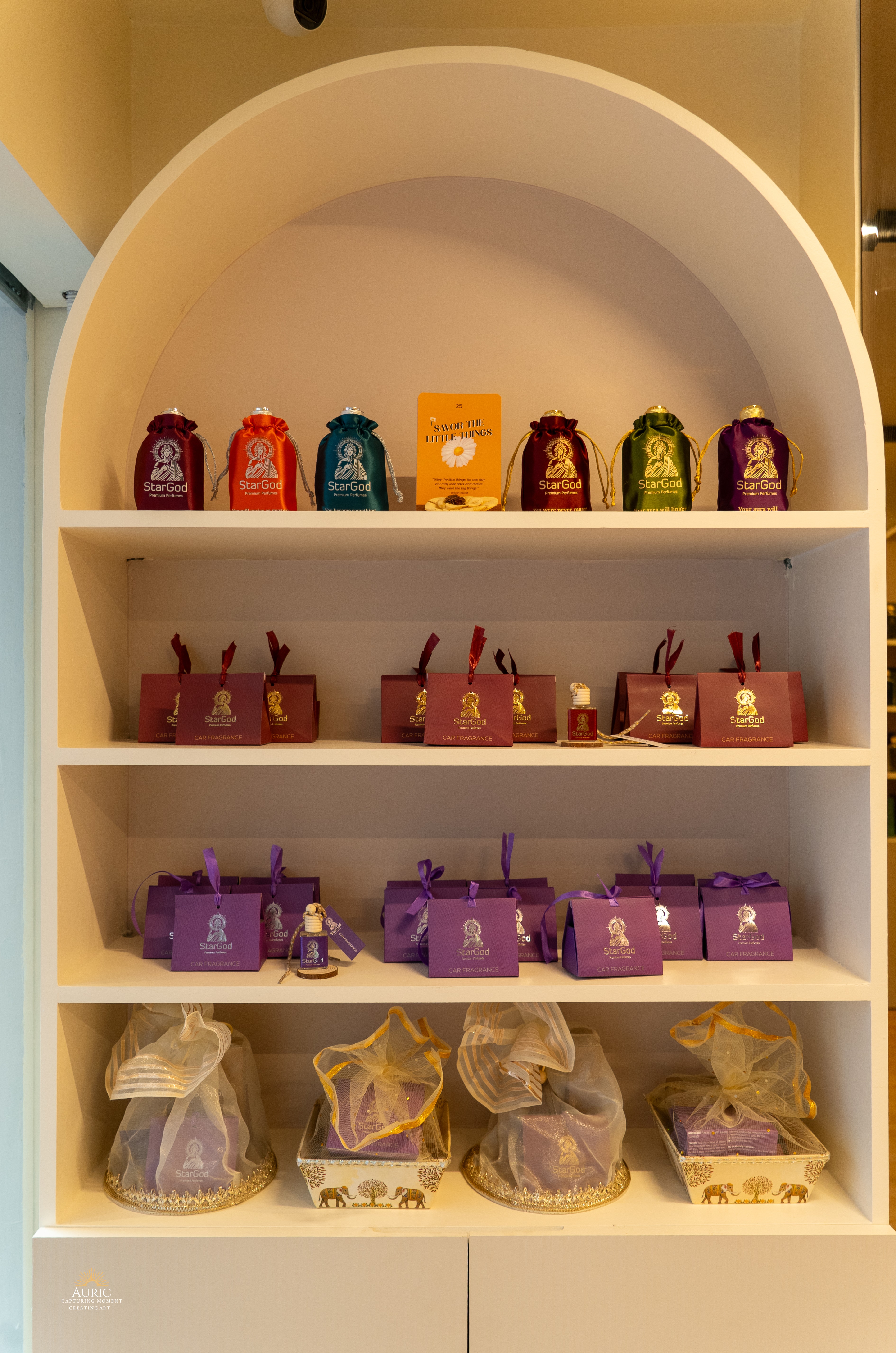 The gifting alcove at the boutique — a rainbow row of satin drawstring pouches on the top shelf, burgundy car-fragrance gift boxes on the second, purple car-fragrance gift boxes on the third, and mesh-wrapped gift hampers in elephant-motif baskets at the bottom.