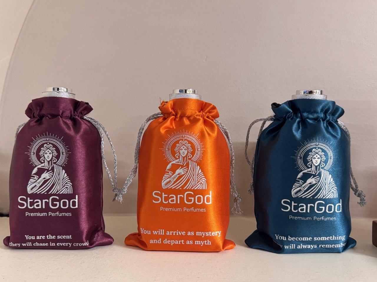 Three StarGod drawstring pouches in aubergine, orange, and teal, each printed with a line in cream.